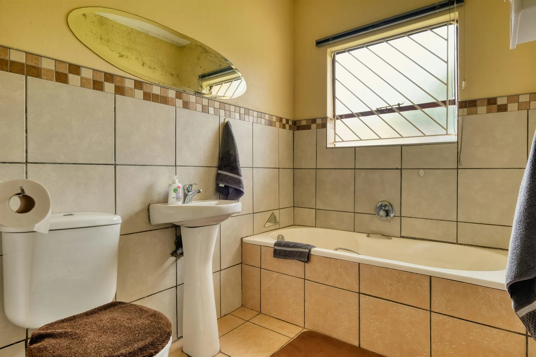 Bathroom with yellow walls, beige tile, tub, pedestal sink, and brighter daylight