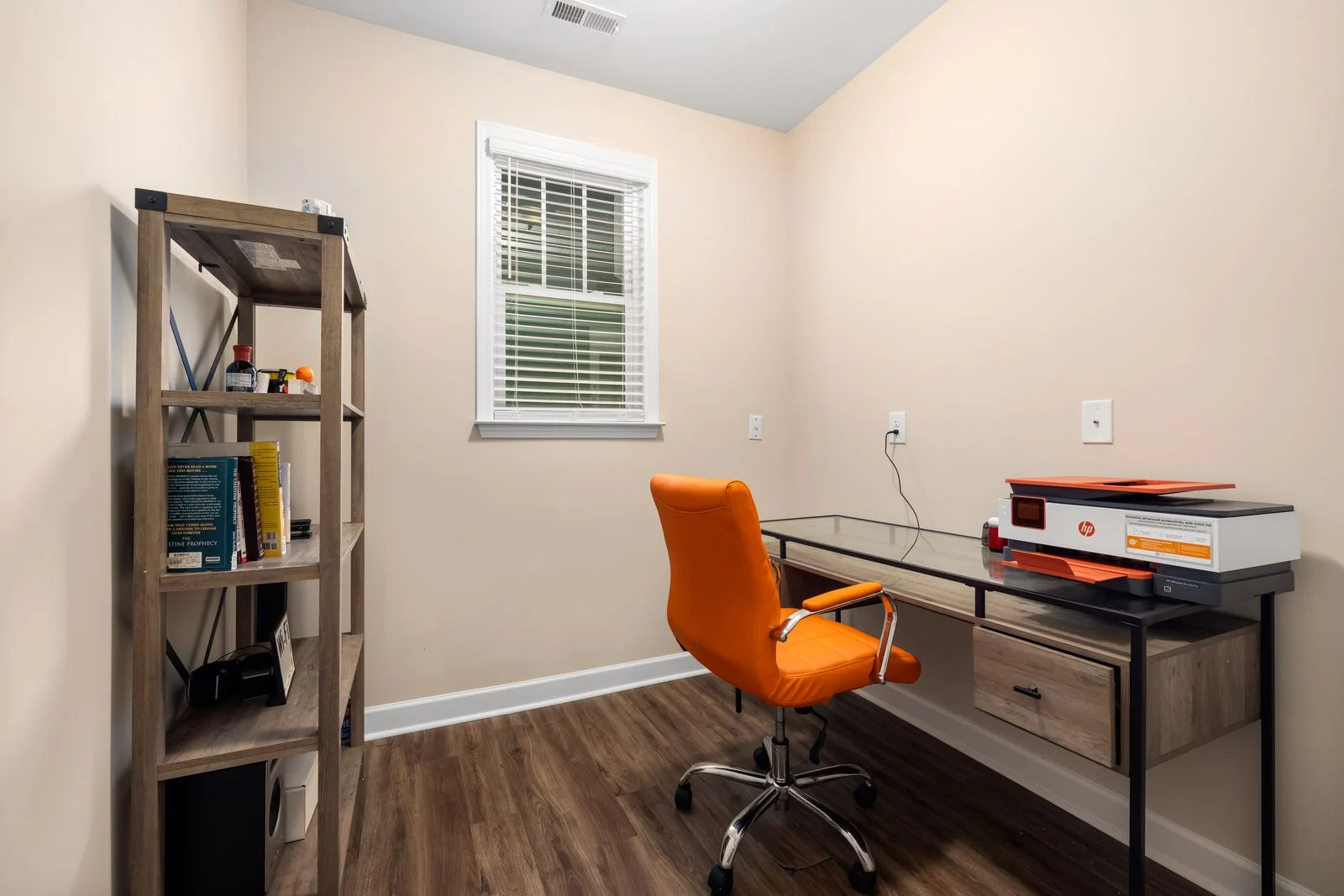 An orange office chair at a wall-facing glass desk with a printer and shelving nearby