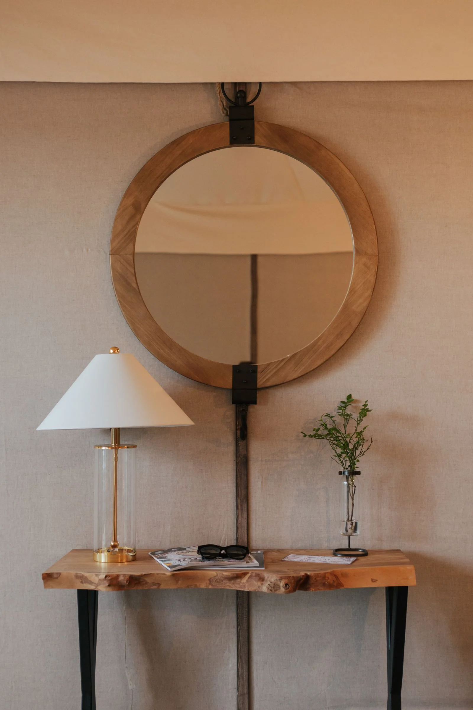 Shaded table lamp on a shallow wooden console beneath a round timber-framed mirror