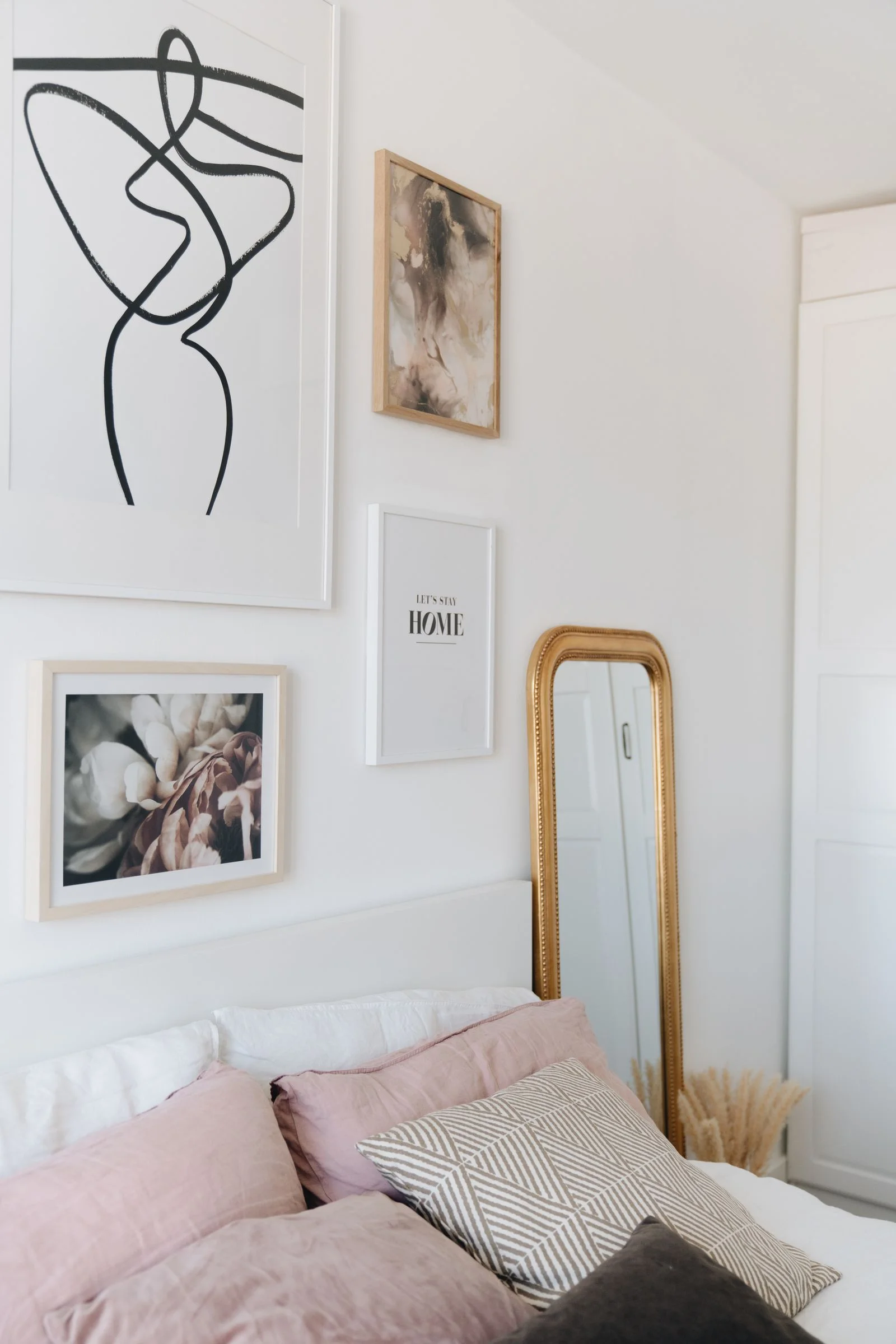 Soft-toned bedroom with floral and abstract framed prints, pale pink cushions and a narrow gold mirror